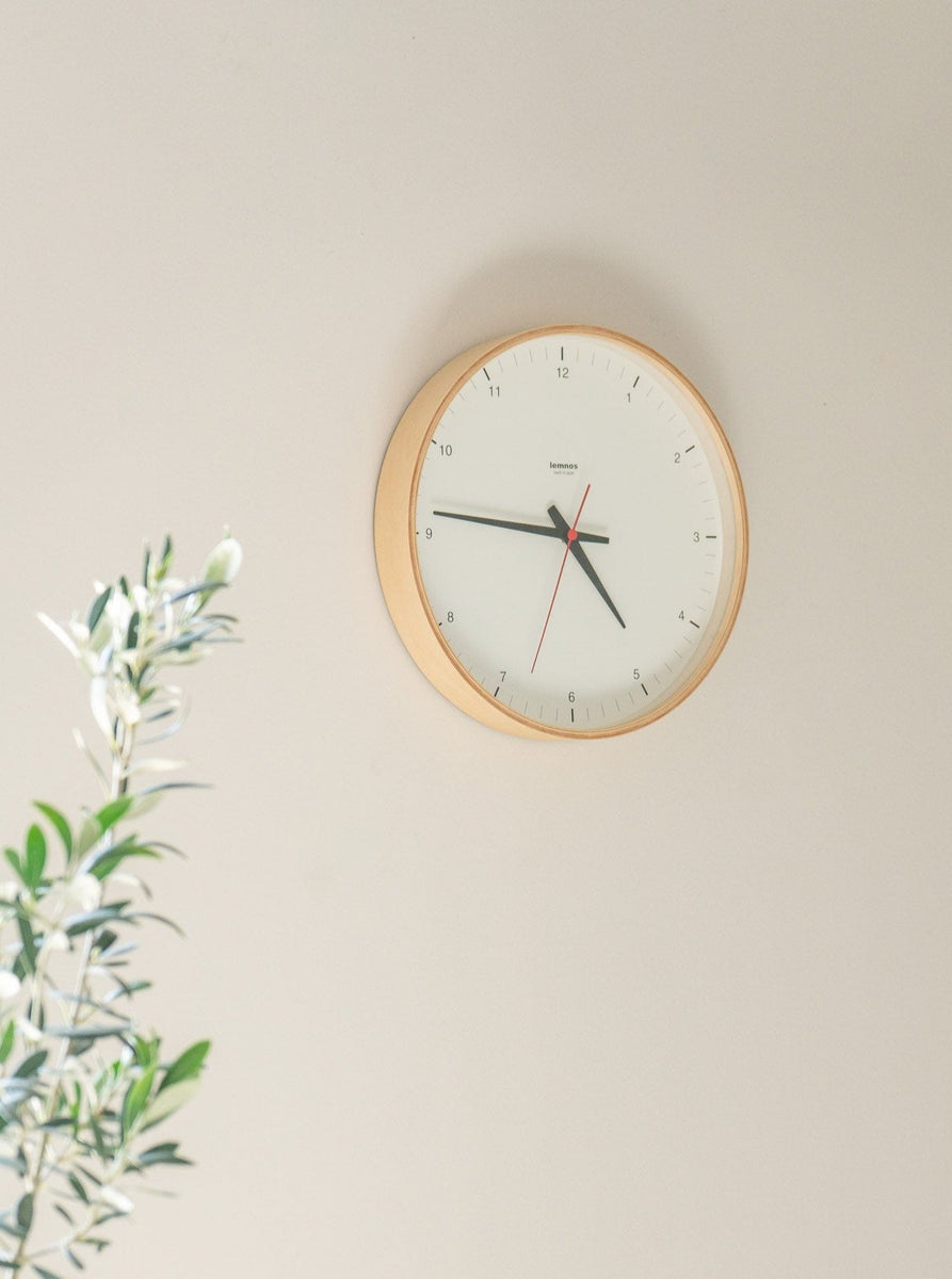 A high-quality Lemnos Plywood Clock handcrafted in Japan, elegantly adorning a wall next to a lush plant.