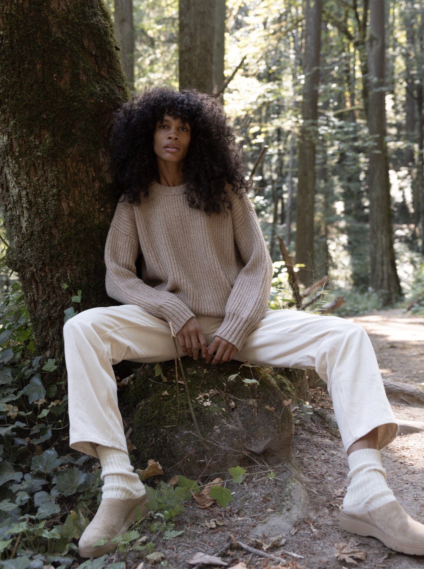 A woman sitting on a rock in the woods wearing a Field Sweater - Caramel and pants.