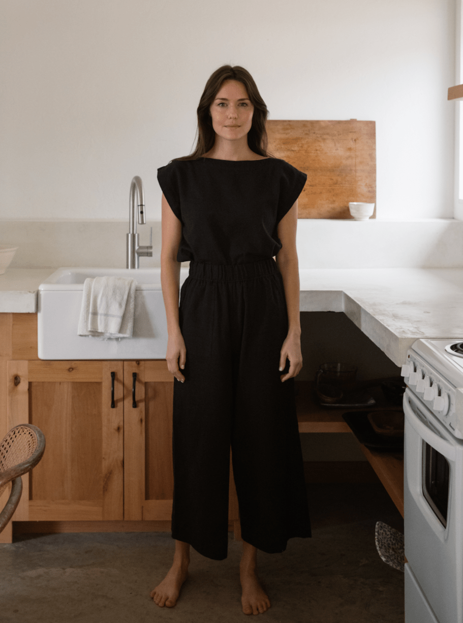 A woman standing in a kitchen wearing the Everyday Crop Pant - Black Cotton.